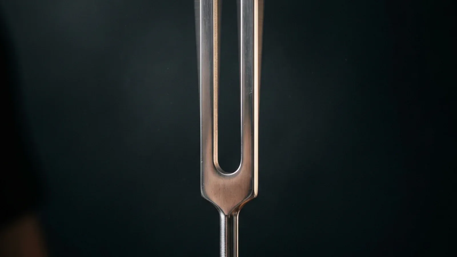 A struck tuning fork held in macro photography against a dark background, with faint motion blur on the tines visualizing real mechanical vibration