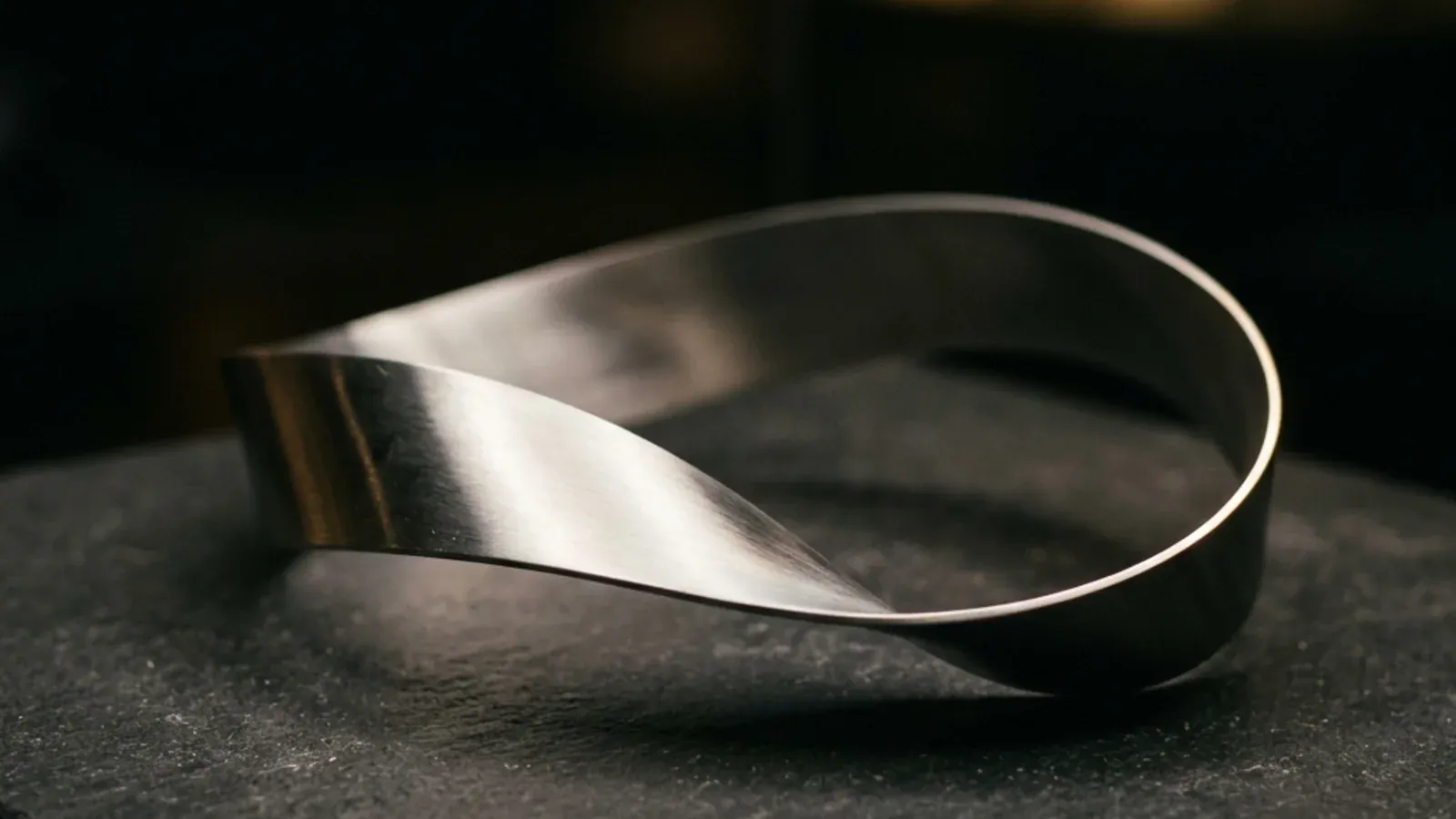A polished metal Möbius strip resting on a dark surface, with soft directional lighting picking out its single continuous edge that you can trace forever without crossing a boundary